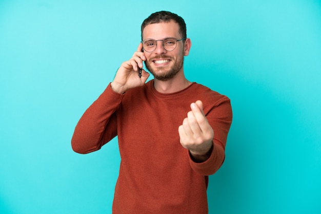 Joven brasileño mediante teléfono móvil aislado sobre fondo azul haciendo gesto de dinero
