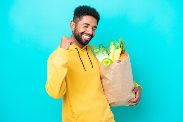 Joven brasileño tomando una bolsa de comida para llevar aislada de fondo azul celebrando una victoria