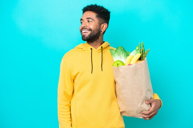 Joven brasileño tomando una bolsa de comida para llevar aislada de fondo azul riéndose en posición lateral