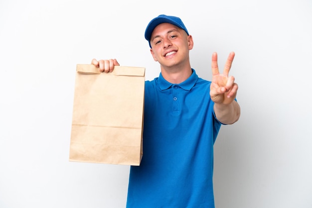 Joven brasileño tomando una bolsa de comida para llevar aislada de fondo blanco sonriendo y mostrando el signo de la victoria