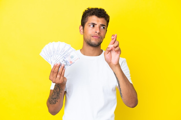 Joven brasileño tomando un montón de dinero aislado sobre fondo amarillo con los dedos cruzando y deseando lo mejor