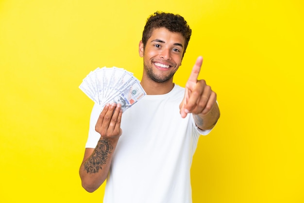 Joven brasileño tomando un montón de dinero aislado sobre fondo amarillo mostrando y levantando un dedo