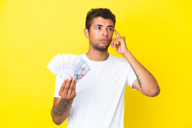 Joven brasileño tomando un montón de dinero aislado sobre fondo amarillo teniendo dudas y pensando