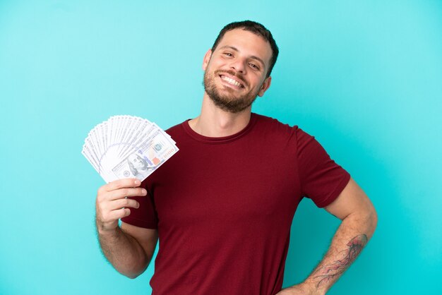 Joven brasileño tomando un montón de dinero sobre antecedentes aislados posando con los brazos en la cadera y sonriendo