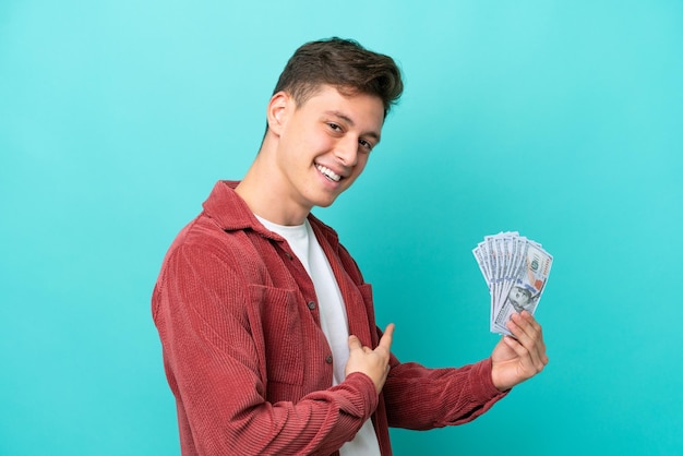 Joven brasileño tomando mucho dinero aislado de fondo azul apuntando hacia atrás