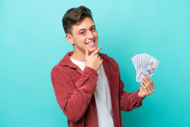 Joven brasileño tomando mucho dinero aislado de fondo azul feliz y sonriente