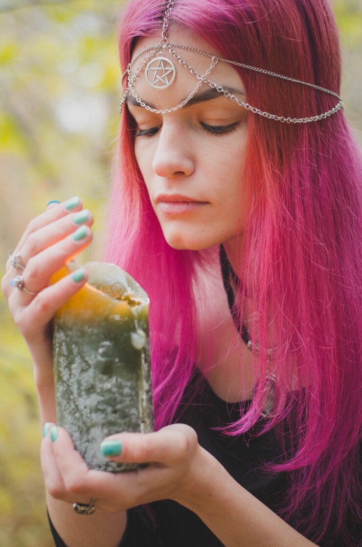 Foto joven bruja con cabello rosado que realiza un ritual mágico con una gran vela verde, colores desteñidos, enfoque seleccionado