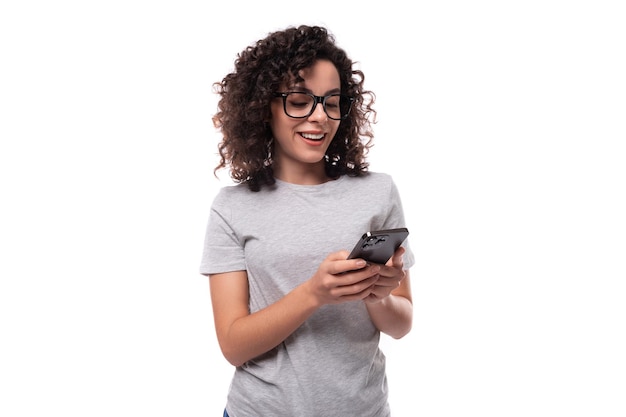 Joven bruneta sonriente mujer rizada vestida con una camiseta gris básica escribe un mensaje en el teléfono