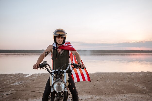 Joven brutal en casco dorado y capa de bandera americana conduciendo motocicleta retro
