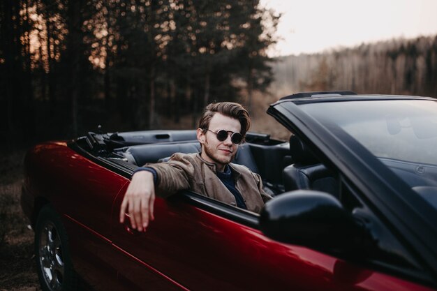 Joven brutal con gafas de sol se sienta en un coche convertible de lujo