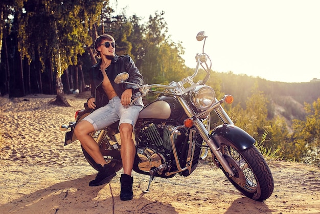 Foto joven brutal con gafas de sol, vaqueros azules y una chaqueta de cuero negro sentado en la costumbre