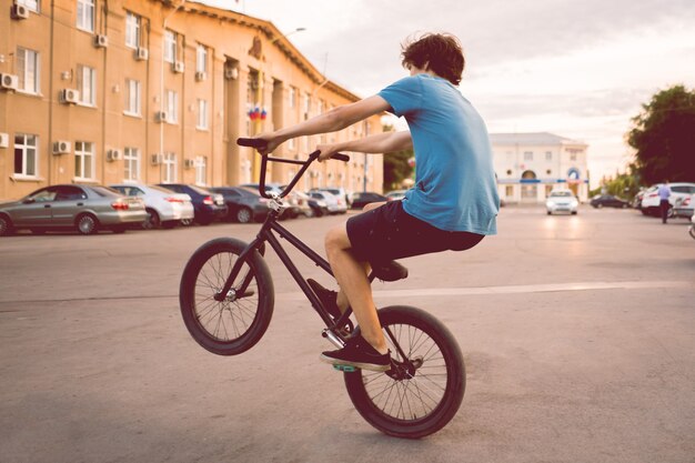 Joven cabalgando sobre una bicicleta bmx en una rueda trasera trasera, haciendo trucos en la ciudad