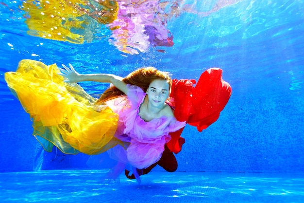 Una joven de cabello largo nada bajo el agua en una piscina al aire libre con telas coloridas en las manos