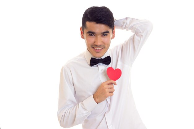 Joven con cabello negro en camiseta blanca con pajarita negra sosteniendo un corazón de papel rojo sobre fondo blanco en el estudio