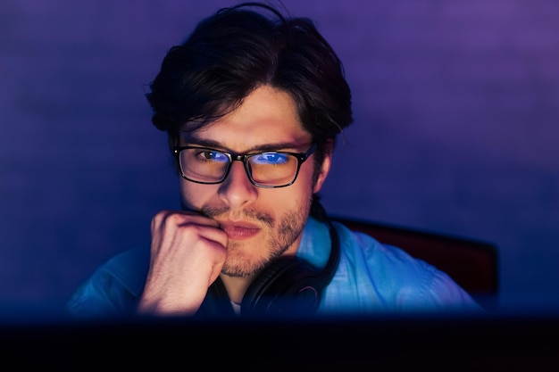 Un joven de cabello oscuro se sienta frente a una pantalla de computadora en una habitación oscura que lleva gafas