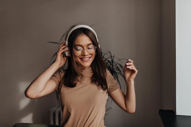 Una joven de cabello oscuro con top beige y anteojos se mueve y sonríe mientras escucha música con grandes auriculares blancos