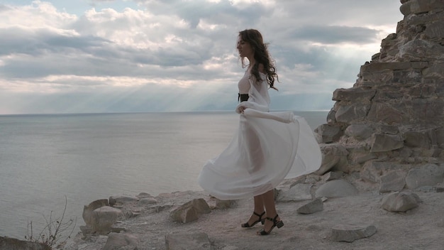 Una joven de cabello oscuro vestida con un vestido blanco está girando en la cima de la montaña el viento soplando su cabello y vestido y sonriendo armonía aventura libertad 4k video cámara lenta