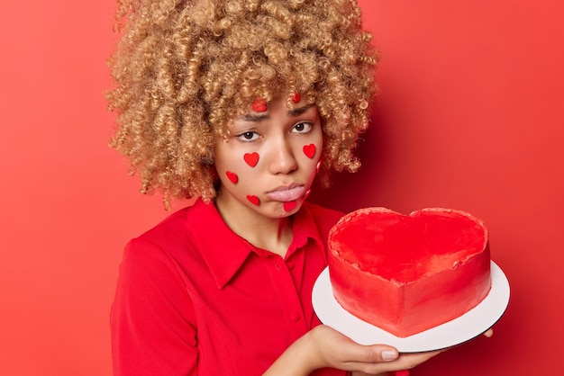 Una joven de cabello rizado disgustada se siente frustrada con una expresión sombría, usa ropa festiva de moda y sostiene un pastel de corazón aislado sobre un fondo rojo que celebra solo el Día de San Valentín