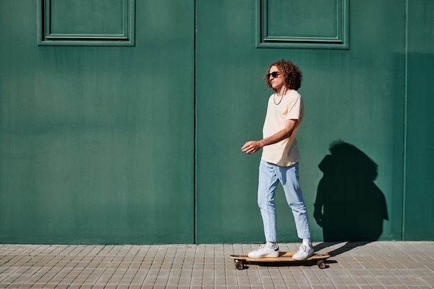 Un joven con cabello rizado y gafas de sol con una pared verde detrás de él en un día soleado