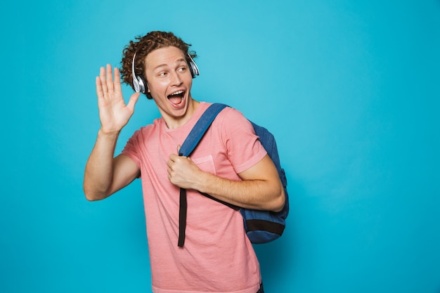 Joven con cabello rizado con mochila escuchando música a través de auriculares y agitando la mano a un lado