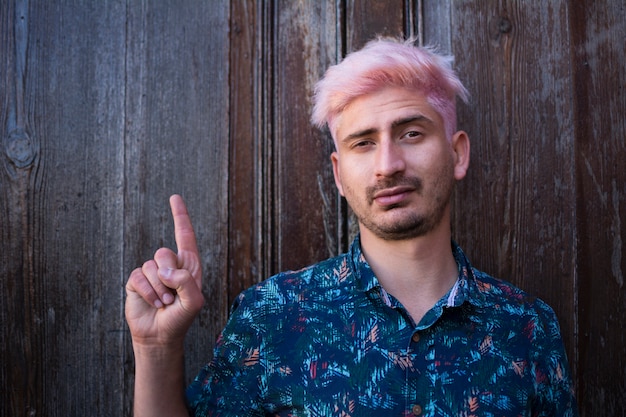 Foto un joven con cabello rosa pastel, vistiendo camisa de verano, apuntando con su dedo, sobre fondo de madera oscura.