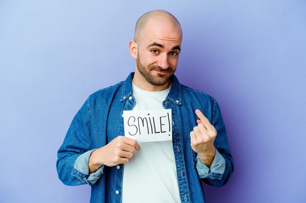 Joven calvo caucásico sosteniendo un cartel de sonrisa aislado en la pared púrpura apuntando con el dedo hacia usted como si estuviera invitando a acercarse