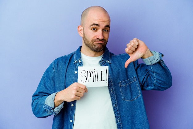 Joven calvo caucásico sosteniendo un cartel de sonrisa aislado en la pared púrpura mostrando un gesto de aversión, pulgares hacia abajo. Concepto de desacuerdo.