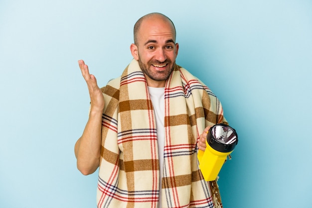 Joven calvo sosteniendo linterna vintage aislado sobre fondo azul recibiendo una agradable sorpresa, emocionado y levantando las manos.