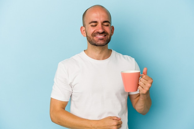 Joven calvo sosteniendo una taza aislada sobre fondo azul riendo y divirtiéndose.