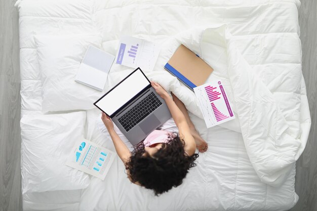 Una joven en la cama con una laptop prepara un informe anual desde casa