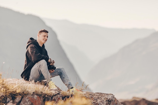 Un joven con una cámara en las manos está sentado en el suelo mirando un maravilloso paisaje montañoso sonriendo. Hay muchas colinas rocosas alrededor.