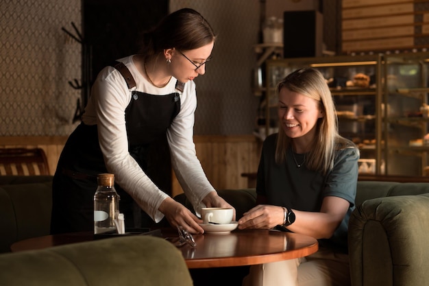 Una joven camarera con delantal sirve café capuchino a una hermosa mujer rubia de negocios en un café