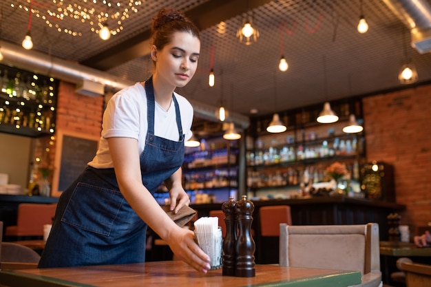 Joven camarera del elegante restaurante poniendo vidrio con un montón de palillos, sal y pimienta en una de las mesas mientras lo prepara para los invitados