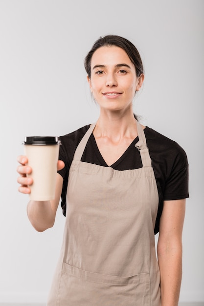 Joven camarera sonriente del café que le da un vaso de papel de café caliente mientras está de pie frente a la cámara en forma aislada