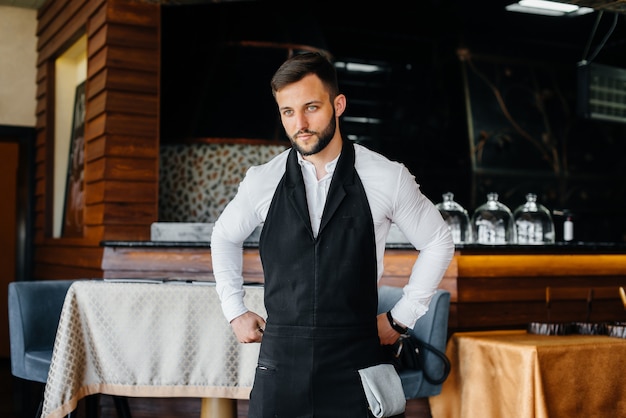 Un joven camarero con barba se pone un delantal y se prepara para una jornada laboral en un buen restaurante.