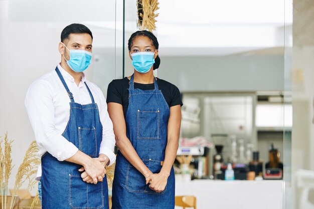 Joven camarero y camarera en máscaras médicas y delantales de pie en la entrada de la pequeña panadería