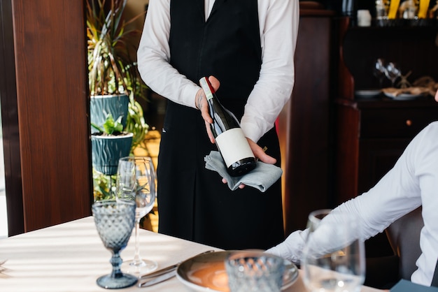 Un joven camarero con un elegante delantal demuestra y ofrece un buen vino a un cliente en un restaurante. Servicio al Cliente.