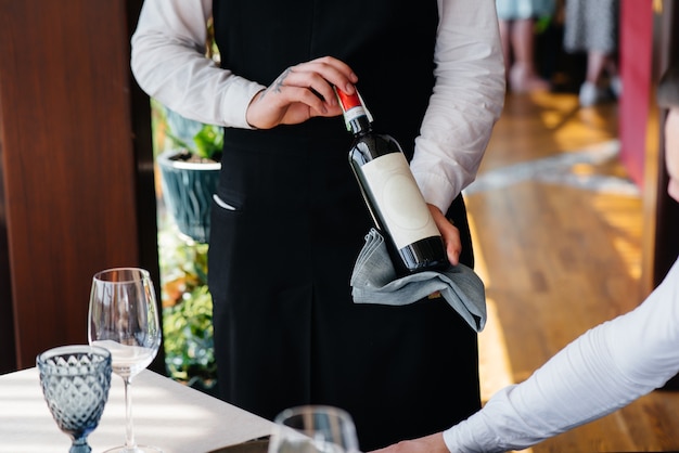 Un joven camarero con un elegante delantal demuestra y ofrece un buen vino a un cliente en un restaurante. Servicio al Cliente.