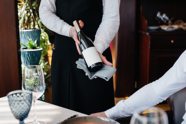 Un joven camarero con un elegante delantal demuestra y ofrece un buen vino a un cliente en un restaurante. Servicio al Cliente.