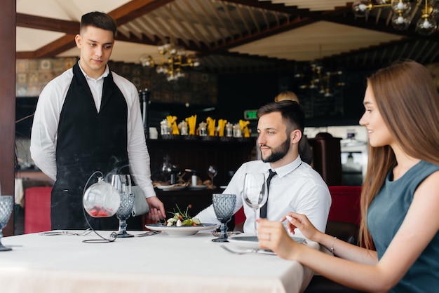 Un joven camarero con un elegante delantal sirve una mesa con una hermosa pareja en un elegante restaurante. Servicio al Cliente.