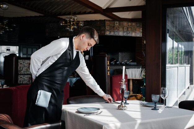 Un joven camarero con un elegante uniforme se dedica a servir la mesa en un hermoso restaurante gourmet Un restaurante de alto nivel Servicio de mesa en el restaurante