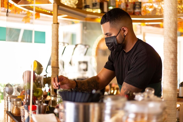 Joven camarero latinoamericano trabajando en el bar. Lleva una máscara de protección facial. Espacio para texto.