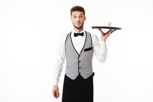 Joven camarero sorprendido en uniforme sosteniendo la bandeja con una taza de café mientras sorprendentemente