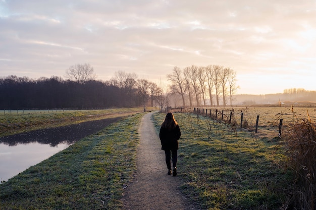 Joven camina en un día frío junto al río en la puesta de sol, fondo hermoso paisaje colorido