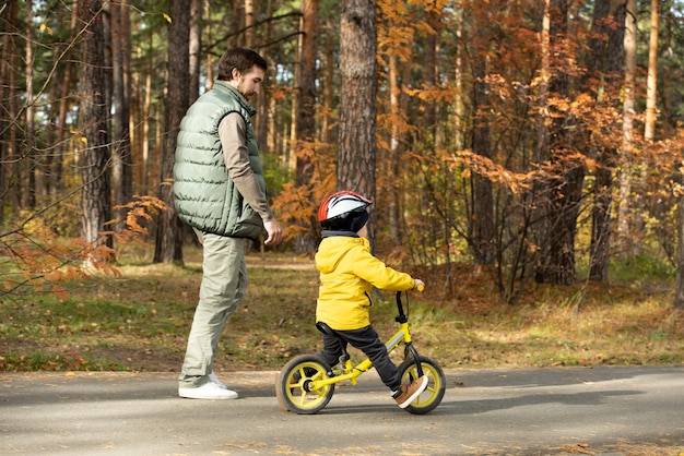 Joven caminando junto a su pequeño hijo en casco de seguridad y ropa casual montando bicicleta de equilibrio a lo largo de la carretera ancha en el parque mientras disfruta de relajarse en el ocio