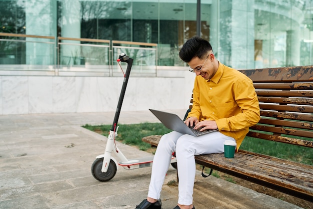 Joven en camisa amarilla estudiando en un parque con laptop y scooter