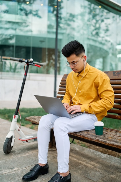 Joven en camisa amarilla estudiando en un parque con laptop y scooter