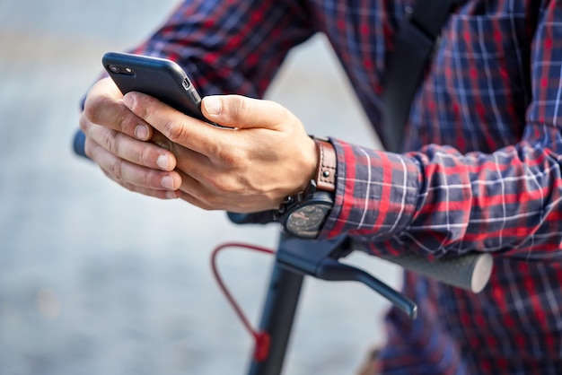 Joven con camisa apoyada en el manillar de scooter eléctrico, sosteniendo un smartphone móvil en sus manos, detalle de cierre.