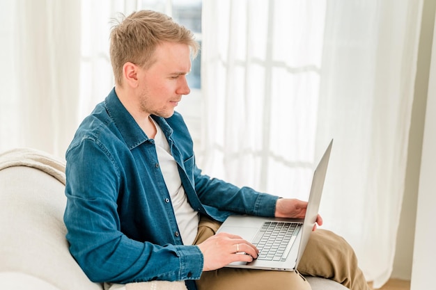 Un joven con una camisa azul está trabajando con una computadora portátil mientras está sentado en el sofá en casa