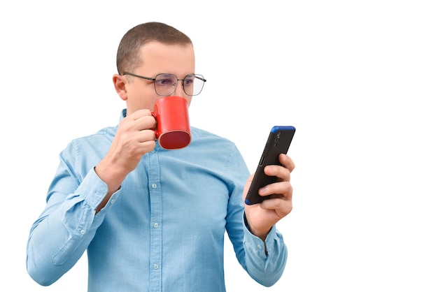 Joven en una camisa bebiendo una bebida caliente y mirando un teléfono móvil sobre un fondo blanco.